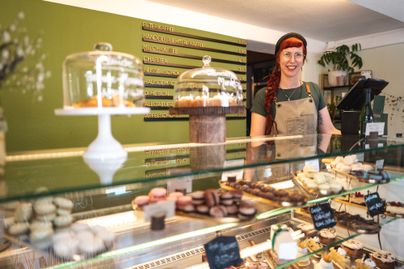 Eine freundliche Frau steht hinter der Theke einer Bäckerei. Vor ihr sind verschiedene, attraktive Gebäckspezialitäten ausgestellt.