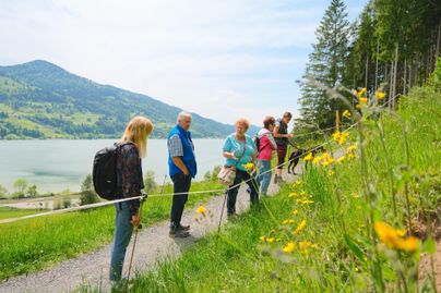 Gruppe von Wanderern, die einen malerischen Weg entlang eines Sees mit Bergen im Hintergrund erkunden. Es blühen gelbe Blumen am Wegesrand.
