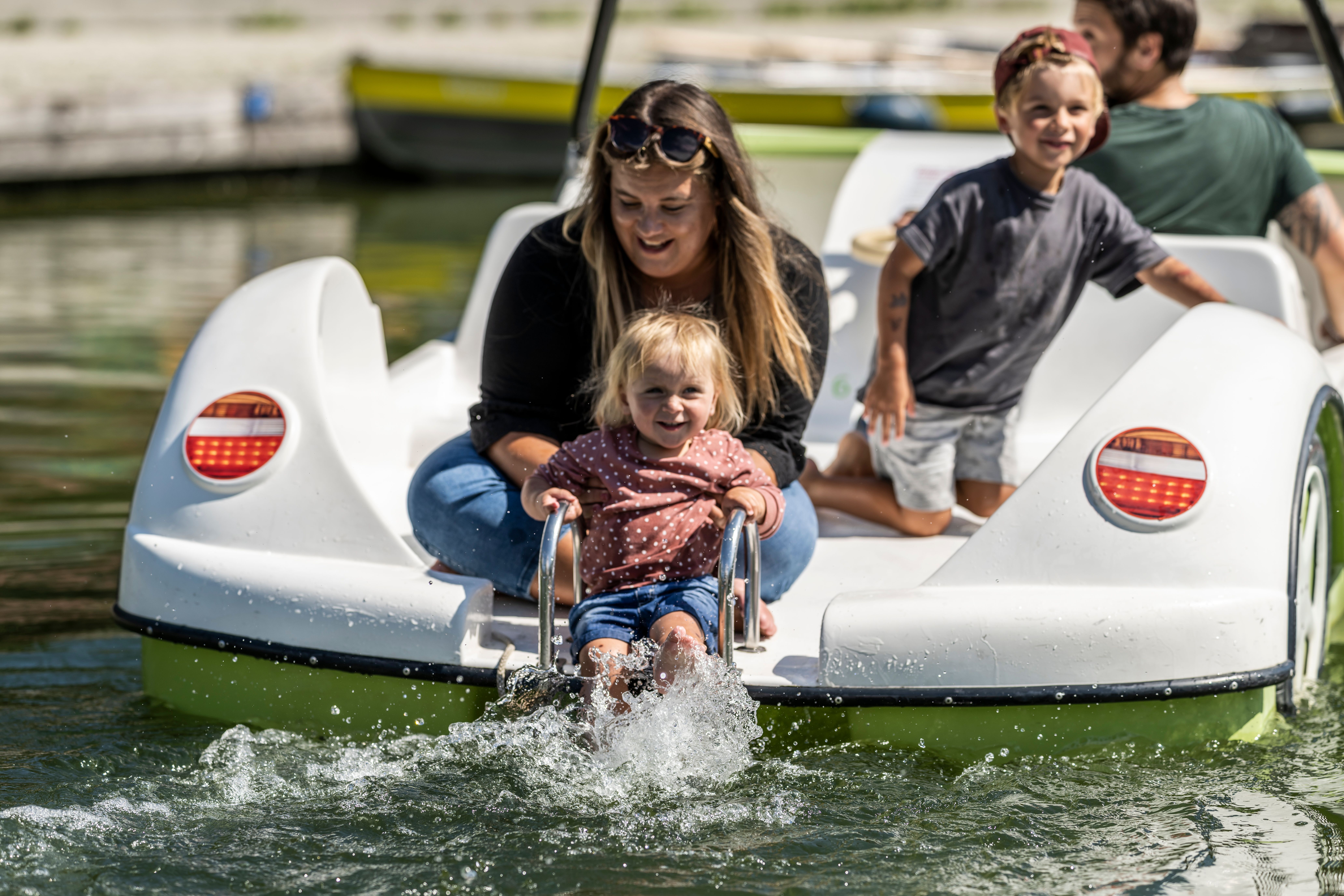 Eine Familie fährt mit einem Tretboot auf dem Alpsee in Immenstadt. Der Papa steuert das Boot, die Mama sitzt mit den beiden Kindern hinten. Das kleine Mädchen paddelt mit den Füßen im Wasser.