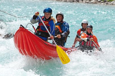 Eine Gruppe von vier Personen paddelt in einem roten Schlauchboot auf einem wilden Fluss. Alle tragen Schwimmwesten und Helme, während sie durch sprudelnde Wellen fahren.