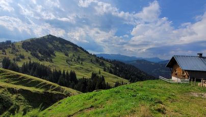 Eine malerische Berglandschaft mit grünen Hügeln und bewaldeten Flächen. Im Vordergrund steht ein traditionelles Holzhaus unter einem klaren Himmel.