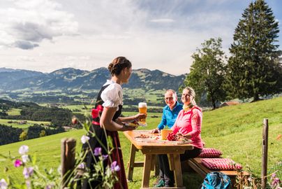 Zwei Wanderer sitzen auf einer Holzbank vor der Bergkulisse. Auf dem Tisch steht eine Brotzeitplatte. Bedienung in Tracht serviert ein Weizenbier.