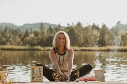 Eine Frau sitzt im Lotussitz am Ufer eines ruhigen Sees. Um sie herum sind spirituelle Statuen und Bilder platziert.