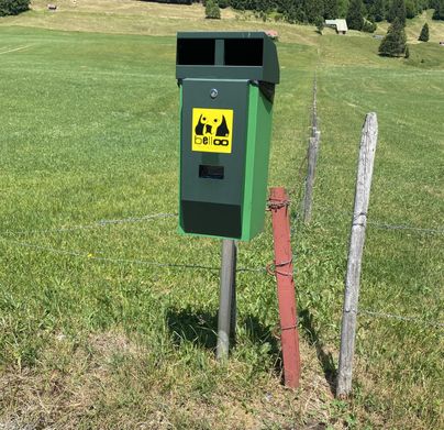 Ein Abfallbehälter steht auf einem grünen Feld. Im Hintergrund sind Bäume und eine Wiese zu sehen.