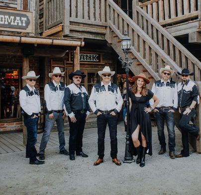 Eine Gruppe von sieben Personen in Western-Kleidung steht vor einem Western-Szenario. Die Männer tragen Cowboyhüte und Hemden, während eine Frau in einem schwarzen Kleid in der Mitte posiert.