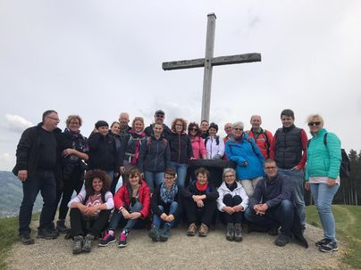 Eine Gruppe von Menschen posiert vor einem großen Kreuz auf einem Hügel. Sie tragen Wanderkleidung und scheinen Freude an ihrem Ausflug zu haben.