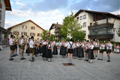 Eine Blaskapelle spielt auf einem Platz, umgeben von traditionellen bayerischen Gebäuden. Die Musiker tragen Trachten und sind in festlicher Stimmung.