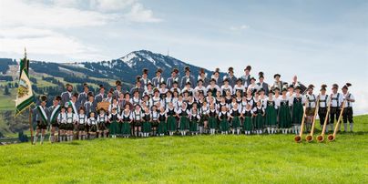 Eine große Gruppe von Menschen in traditioneller Tracht steht auf einer grünen Wiese. Im Hintergrund sind Berge und ein klarer Himmel zu sehen.