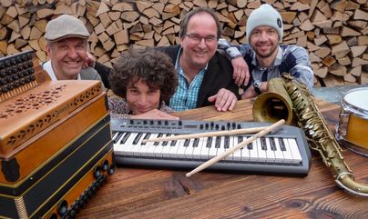 Eine Gruppe von vier Musikern posiert vor einem Haufen Holzscheite. Sie haben verschiedene Musikinstrumente wie ein Akkordeon, eine Klavier-Tastatur und ein Saxophon dabei.