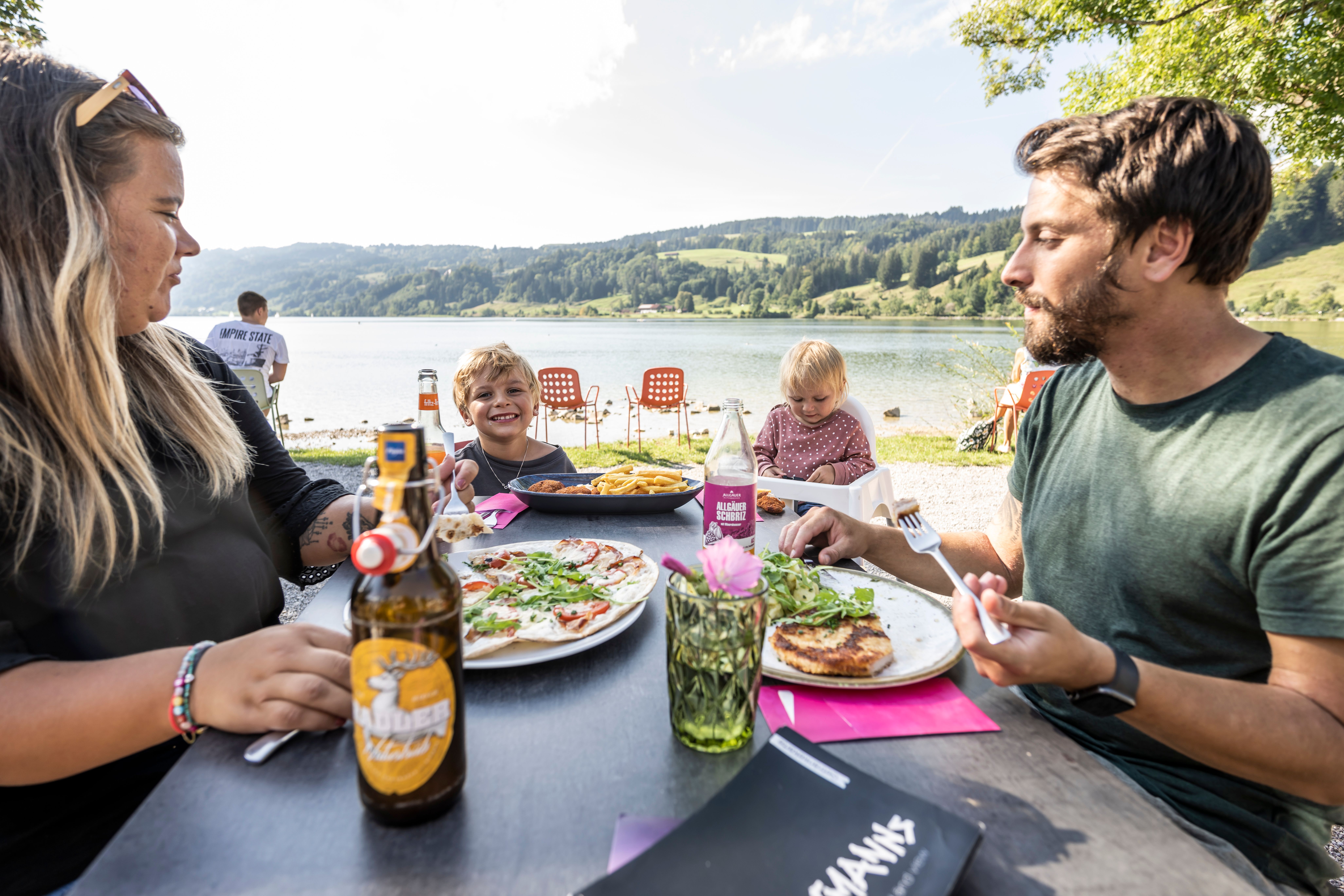 Eine Familie mit Mama, Papa und zwei Kindern sitz an einem Tisch verzehrt verschiedene Speisen und Getränke. Im Hintergrund ist der Alpsee in Immenstadt zu sehen.