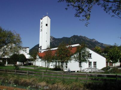 Ein modernes Gebäude mit einem schlanken Turm steht in einer ländlichen Umgebung. Im Hintergrund sind grüne Berge und ein klarer blauer Himmel sichtbar.