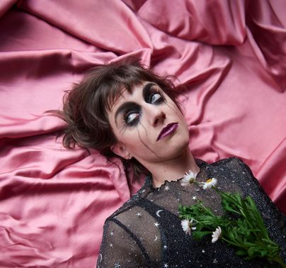 Eine Person liegt auf einem glänzenden pinken Tuch und trägt ein glitzerndes schwarzes Outfit. Sie hat auffälliges Makeup und hält eine kleine Blume in der Hand.