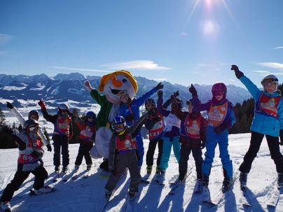 Eine Gruppe fröhlicher Kinder posiert im Schnee, umgeben von Bergen. Sie tragen Skiausrüstung und haben Spaß mit einem Maskottchen.