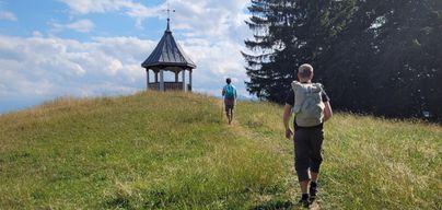 Zwei Personen gehen auf einem grasbewachsenen Hügel in Richtung eines Pavillons. Im Hintergrund sind Bäume und ein klarer Himmel zu sehen.