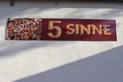 Ein Schild mit der Aufschrift „5 SINNE“ und einem Bild von kleinen Kieselsteinen. Der Hintergrund ist hell und neutral.
