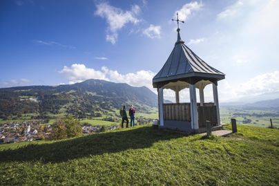 Eine Aussichtsplattform auf einem Hügel mit Blick auf eine grüne Landschaft und Berge im Hintergrund. Zwei Personen stehen vor der Plattform und genießen die Aussicht.