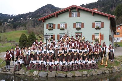 Eine große Musikkapelle steht vor einem grünen Gebäude in einer ländlichen Umgebung. Die Mitglieder tragen traditionelle Trachten und halten verschiedene Instrumente in der Hand.