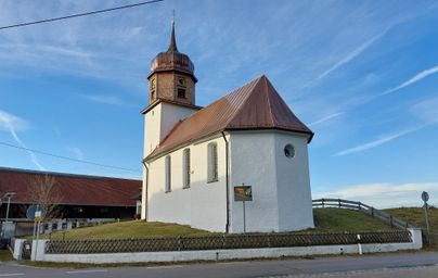 Eine kleine Kirche mit einem hohen Turm und einem schrägen Dach. Sie steht auf einem Hügel im Freien unter einem klaren Himmel.