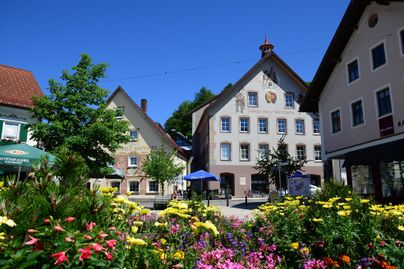 Ein malerischer Platz mit bunten Blumenbeeten und historischen Gebäuden. Der Himmel ist klar und blau, was eine angenehme Atmosphäre schafft.