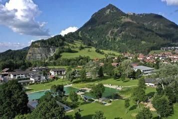 Eine malerische Landschaft mit einem großen grünen Park und einem Berg im Hintergrund. Im Vordergrund sind Schwimmbecken und Bäume zu sehen.