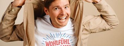 Ein fröhlicher Mann zieht seine Jacke aus und zeigt ein T-Shirt mit der Aufschrift "Wohlfühl". Er lächelt und strahlt Energie aus.