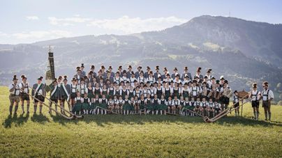 Eine große Gruppe von Menschen in traditioneller Tracht steht auf einer Wiese in den Bergen. Im Hintergrund sind grüne Hügel und ein klarer Himmel zu sehen.