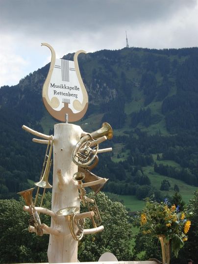 Ein Aushang der Musikgruppe "Musikkapelle Rettenberg" mit verschiedenen Musikinstrumenten. Im Hintergrund sieht man eine grüne Berglandschaft unter einem bewölkten Himmel.