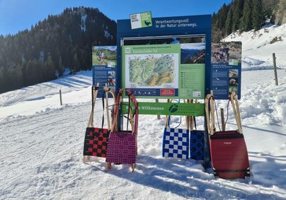 Eine Anmeldungstafel im Schnee mit einer Karte der Umgebung. Im Vordergrund hängen bunte Rodelschlitten.