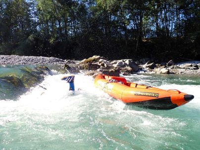 Ein orangefarbenes Schlauchboot kämpft gegen die Wellen in einem Fluss. Im Hintergrund sind Bäume und Felsen sichtbar.
