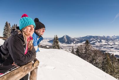 Ein Mann und eine Frau blicken im Winter, gelehnt an einen Zaun, vom Gipfel des Ofterschwanger Horns in die Ferne. Das 1.406 Meter hohe Ofterschwanger Horn erhebt sich über der Gemeinde Ofterschwang und liegt südlich von Gunzesried (Blaichach).