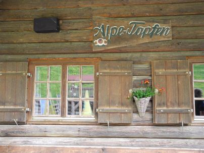 Ein traditionelles Holzhaus mit einem Schild "Alpe-Tropfen". Die Fensterläden sind geschlossen und es gibt einen Blumenkasten mit Blumen.