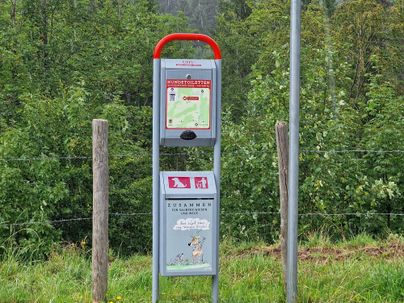 Eine Informationsstelle mit Karten und Wachhund-Symbolen steht in der Natur. Im Hintergrund sind grüne Bäume sichtbar.