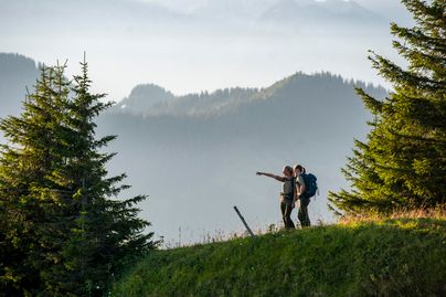 Ein Paar steht auf einem Hügel und schaut in die Ferne. Im Hintergrund sind malerische Berge und grüne Bäume zu sehen.