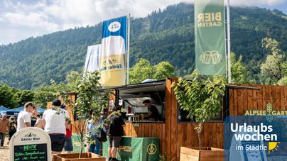 Ein belebter Außenbereich mit einem Holzstand und Menschen, die anstehen. Hintergrund ist eine grüne Landschaft und ein blauer Himmel.