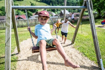 Ein fröhliches Kind schwingt auf einer Schaukel. Im Hintergrund spielen zwei weitere Kinder auf einem sonnigen Spielplatz.