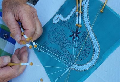 Hände arbeiten an einem feinen Spitzenprojekt auf einem blauen Stoff. Die verwendeten Werkzeuge sind Nadel und Faden.