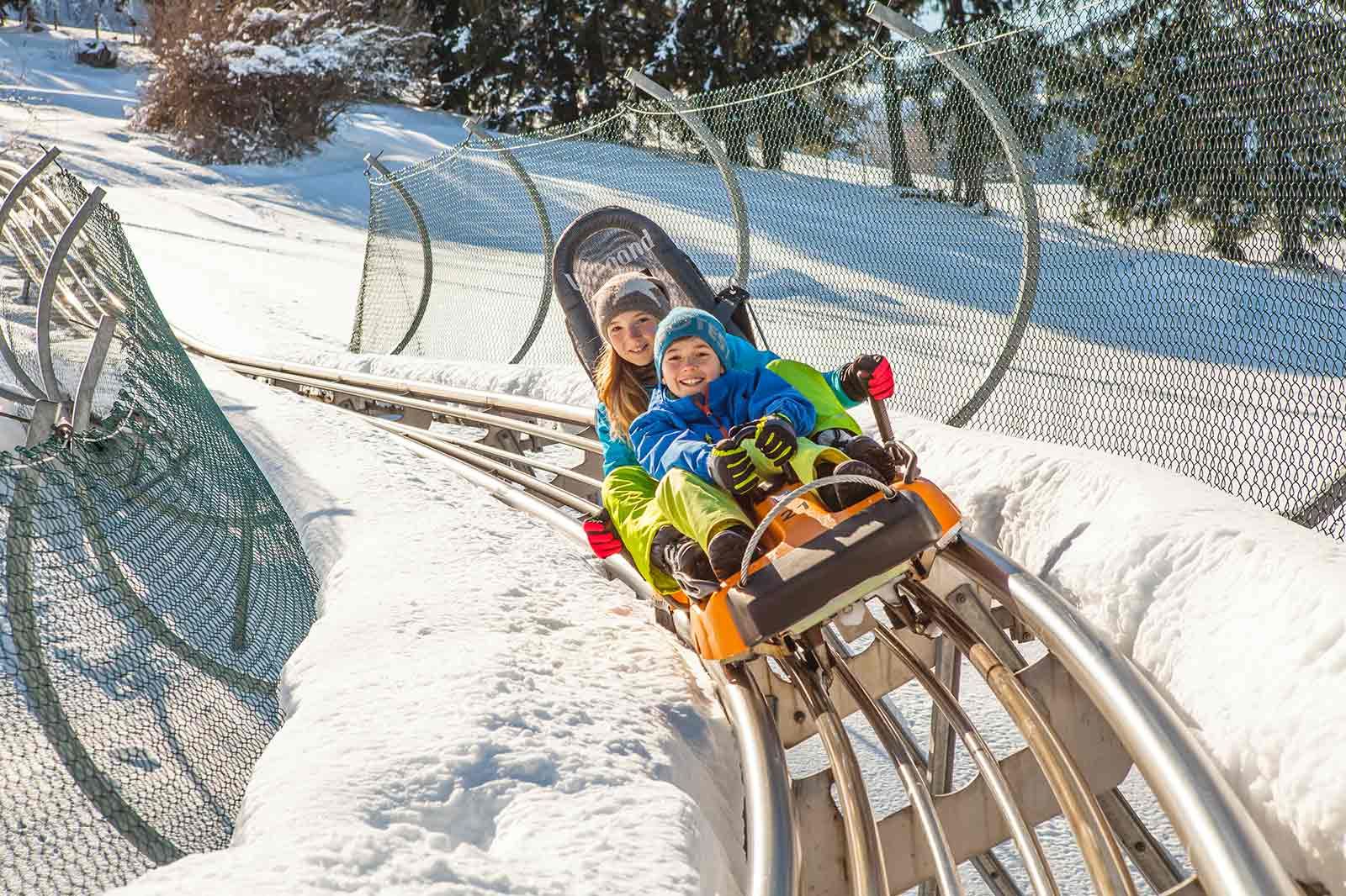 Zwei Kindern rodeln mit dem Alpsee Coaster der Alpsee Bergwelt bei Immenstadt im Winter den Berg hinunter.