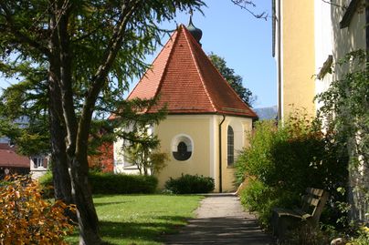 Eine kleine Kapelle mit einem roten Dach steht in einem grünen Garten. Umgeben von Bäumen und blühenden Pflanzen, strahlt die Szene Ruhe und Harmonie aus.