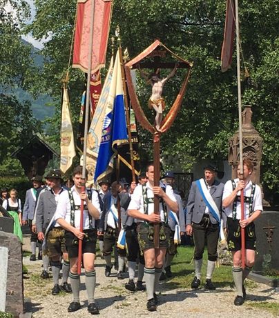 Eine Gruppe von Festteilnehmern trägt Fahnen und Schreine bei einem traditionellen Umzug. Sie tragen Trachten und bewegen sich durch eine malerische Umgebung.