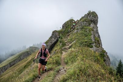 Eine Frau joggt fröhlich auf einem schmalen Pfad in den Bergen. Im Hintergrund sind graue Wolken und eine grüne Landschaft zu sehen.