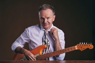 Ein Mann sitzt mit einer E-Gitarre in der Hand. Er trägt ein Hemd und eine Krawatte mit auffälligem Muster.