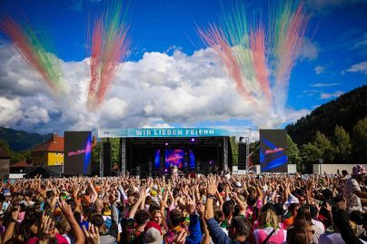 Eine lebhafte Festivalatmosphäre mit einer großen Menschenmenge vor einer Bühne. Farbenfrohe Feuerwerke erleuchten den Himmel bei strahlendem Wetter.
