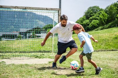 Ein Mann spielt mit einem Jungen Fußball auf einem grünen Platz. Im Hintergrund ist ein Fußballtor und eine hügelige Landschaft zu sehen.