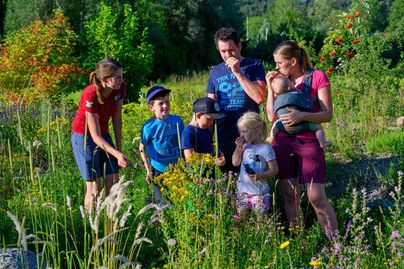 Eine Gruppe von Kindern und Erwachsenen steht in einem bunten Garten mit vielen Blumen. Sie scheinen die Pflanzen zu erkunden und zu genießen.
