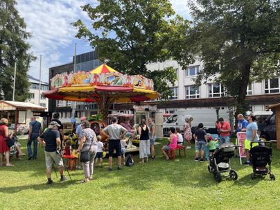 Ein bunter Jahrmarkt mit einem Karussell und mehreren Attraktionen. Viele Menschen genießen die festliche Atmosphäre auf der Wiese.