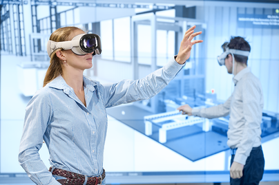 Eine Frau mit einer Virtual-Reality-Brille interagiert mit einer digitalen Umgebung. Im Hintergrund ist ein Mann ebenfalls mit VR-Brille zu sehen, der an einem interaktiven Bildschirm arbeitet.