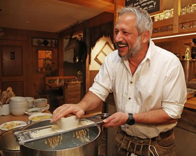 Ein lächelnder Mann reibt Käse über einen großen Topf in einem rustikalen Restaurant. Der Hintergrund zeigt eine gemütliche Atmosphäre mit Holzverzierungen.