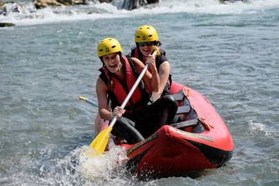 Zwei Frauen paddeln in einem roten Schlauchboot auf einem Fluss. Beide tragen gelbe Helme und haben Spaß.