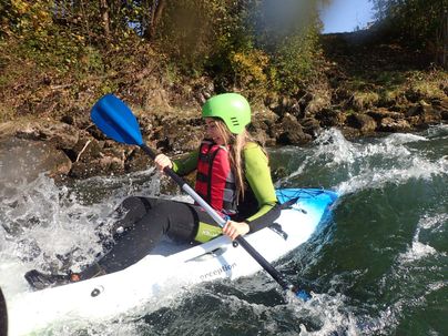 Eine Person paddelt in einem Kajak durch strömendes Wasser. Sie trägt einen grünen Helm und eine Schwimmweste.