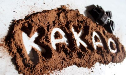 Kakaopulver liegt verstreut auf einer Oberfläche, und das Wort „Kakao“ ist in das Pulver geschrieben. Daneben befinden sich einige Stücke Schokolade.
